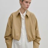 A person wearing a relaxed fit, cropped tan Stella Jacket by Herskind ApS over a long-sleeved white button-down shirt.