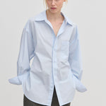 A model wears an oversized cotton Jordan Shirt by Herskind ApS with a point collar, breast pocket, and cuffed sleeves, tucked into dark grey trousers.