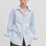 A model wears an oversized cotton Jordan Shirt by Herskind ApS with a point collar, breast pocket, and cuffed sleeves, tucked into dark grey trousers.