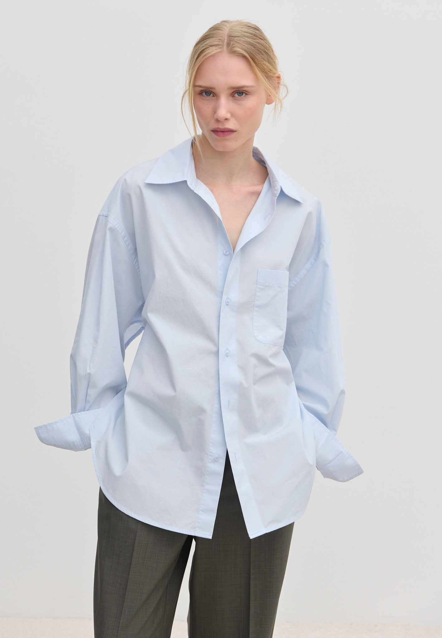A model wears an oversized cotton Jordan Shirt by Herskind ApS with a point collar, breast pocket, and cuffed sleeves, tucked into dark grey trousers.