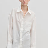 A person wears the oversized, long-sleeved white Jordan Shirt by Herskind ApS, featuring a breast pocket and a curved hem.