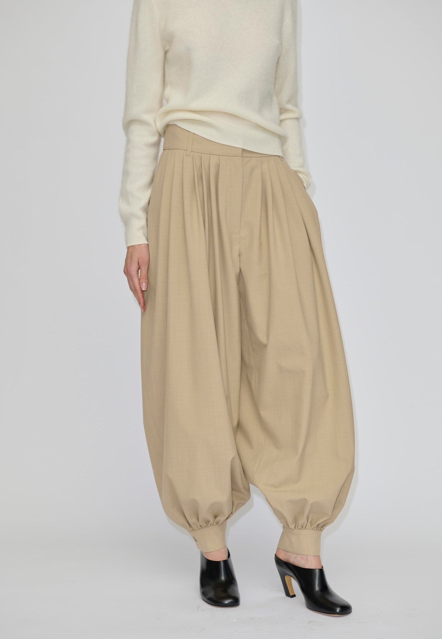 Herskind ApS Zeth High Waist Pants in khaki, featuring a high-waisted, pleated design with a relaxed fit and tapered, cuffed hem, worn with a cream sweater and black heeled mules.