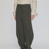 Person wearing a beige long-sleeved button-down shirt tucked into mid-waist, olive Dante Mid Waist Pants by Herskind ApS featuring a barrel leg shape with a slight taper at the hem and light-colored shoes.