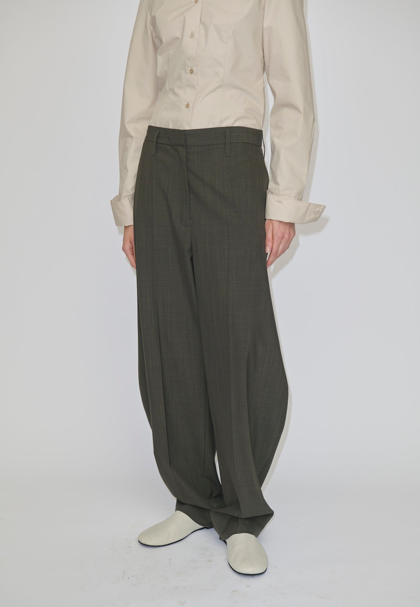 Person wearing a beige long-sleeved button-down shirt tucked into mid-waist, olive Dante Mid Waist Pants by Herskind ApS featuring a barrel leg shape with a slight taper at the hem and light-colored shoes.
