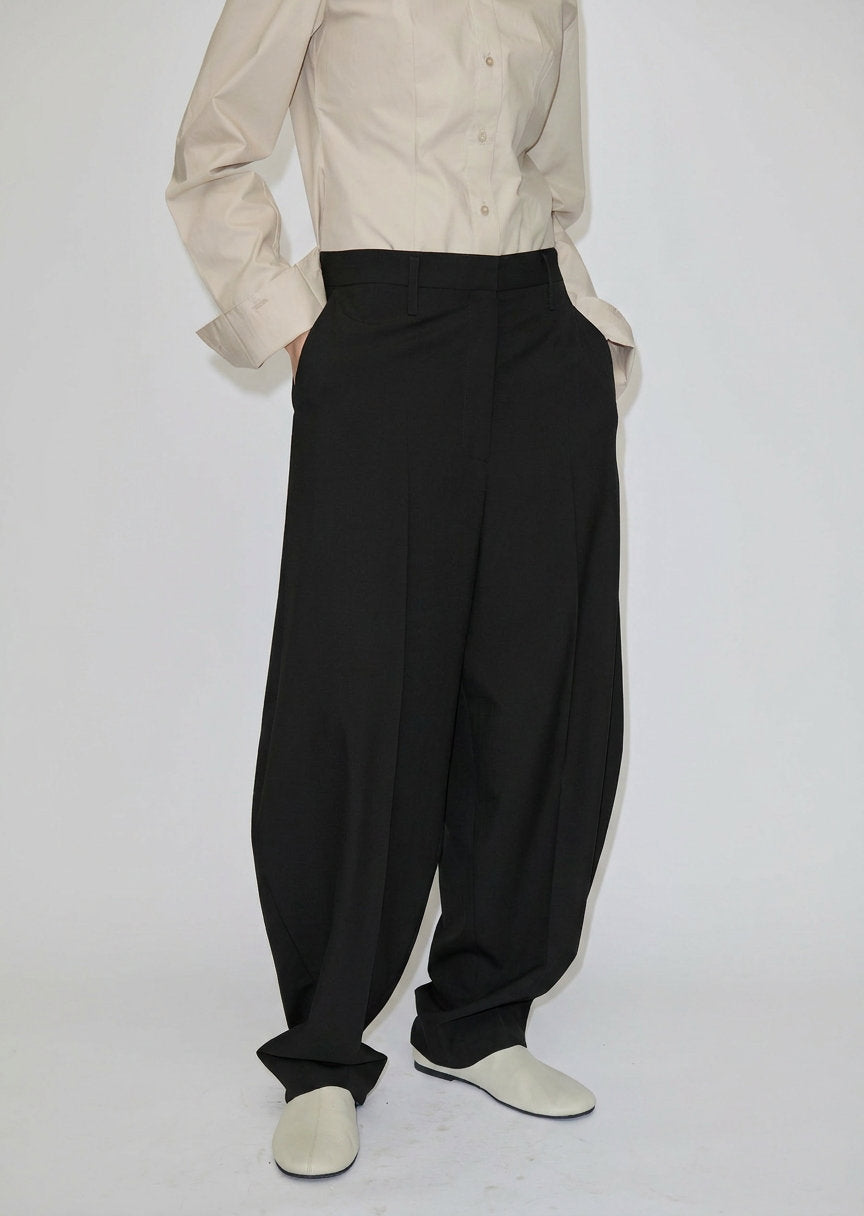 A person wearing a beige button-down shirt and black Dante Mid Waist Pants, featuring a high-waisted, relaxed barrel leg fit with front pleats.