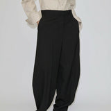 A person wearing a beige button-down shirt and black Dante Mid Waist Pants, featuring a high-waisted, relaxed barrel leg fit with front pleats.