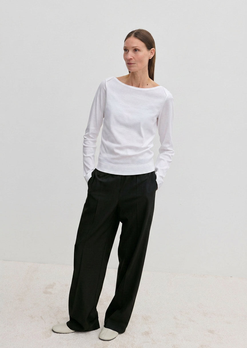 A woman wears a white long-sleeved shirt and Pinky Mid waist Pants in black with an elasticated waistband, her hands tucked into her pockets.