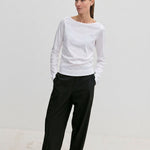 A woman wears a white long-sleeved shirt and Pinky Mid waist Pants in black with an elasticated waistband, her hands tucked into her pockets.