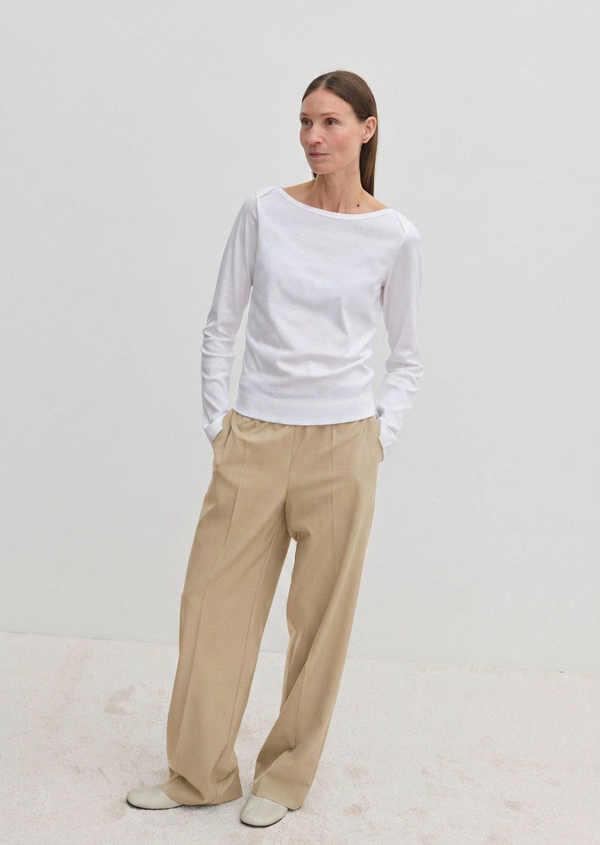A woman wears a white long-sleeved shirt and Pinky Mid waist Pants, which are wide-leg beige trousers with an elasticated waistband.