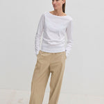 A woman wears a white long-sleeved shirt and Pinky Mid waist Pants, which are wide-leg beige trousers with an elasticated waistband.