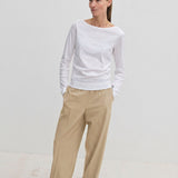 A woman wears a white long-sleeved shirt and Pinky Mid waist Pants, which are wide-leg beige trousers with an elasticated waistband.