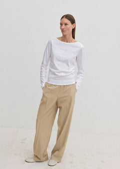 A woman wears a white long-sleeved shirt and Pinky Mid waist Pants, which are wide-leg beige trousers with an elasticated waistband.