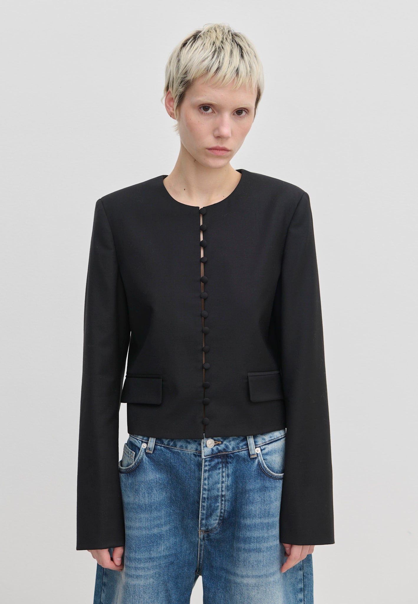 Herskind ApS Marge Fitted Blazer: a black collarless cropped blazer with a front row of fabric-covered buttons, worn over blue jeans.