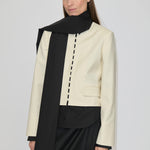 A person wears a cream-colored, collarless cropped Marge Fitted Blazer by Herskind ApS with a row of small buttons down the front, layered over a black shirt and a long black scarf.