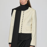 A person wears a cream-colored, collarless cropped Marge Fitted Blazer by Herskind ApS with a row of small buttons down the front, layered over a black shirt and a long black scarf.