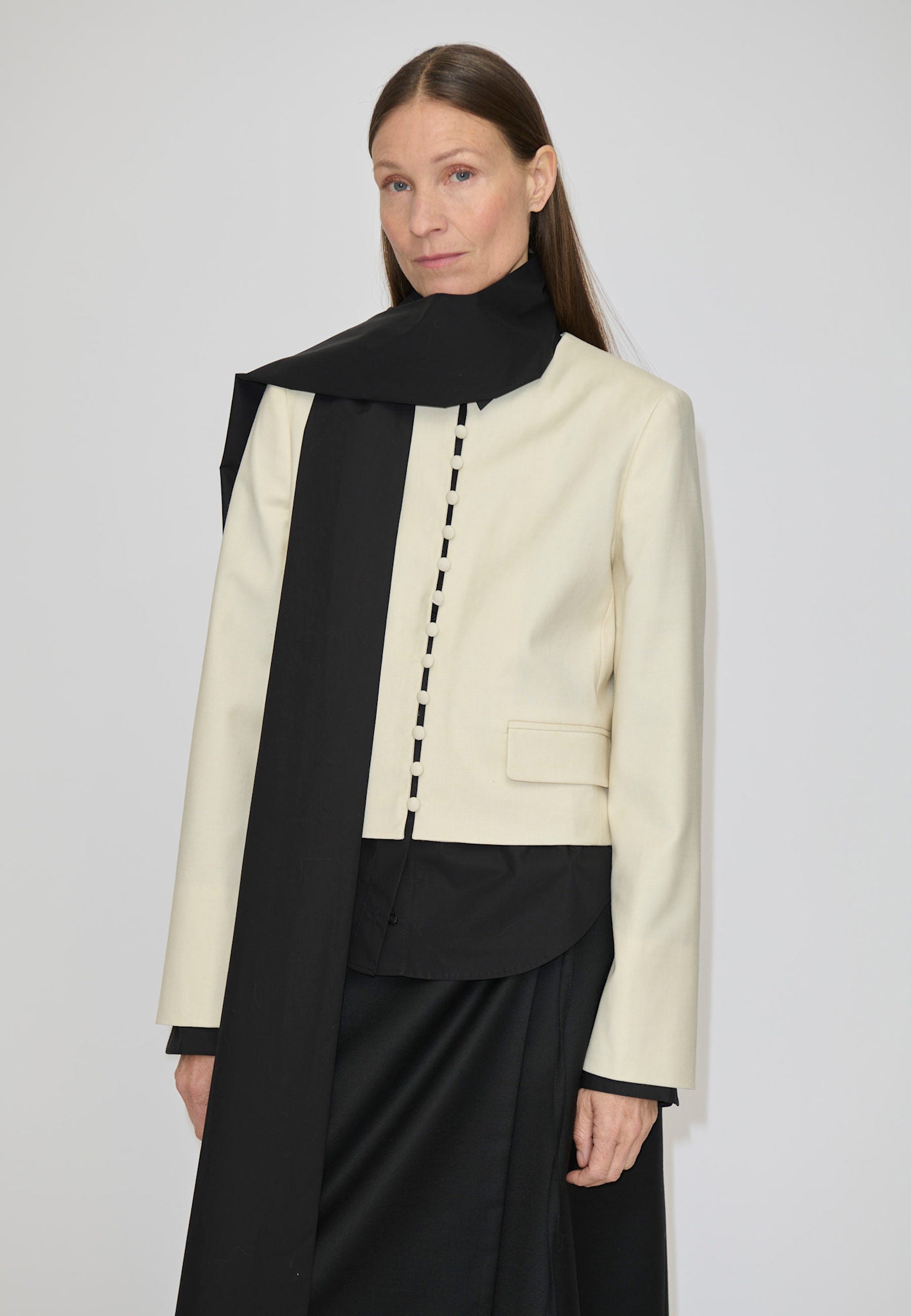 A person wears a cream-colored, collarless cropped Marge Fitted Blazer by Herskind ApS with a row of small buttons down the front, layered over a black shirt and a long black scarf.