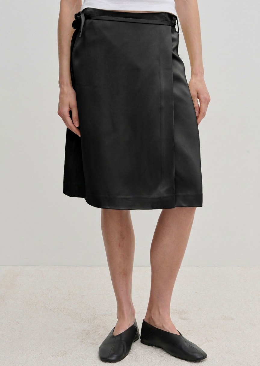 A person wearing a black knee-length Herskind ApS Emmy Wrap Skirt tied at the waist with black slip-on shoes.