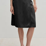 A person wearing a black knee-length Herskind ApS Emmy Wrap Skirt tied at the waist with black slip-on shoes.