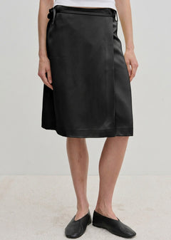 A person wearing a black knee-length Herskind ApS Emmy Wrap Skirt tied at the waist with black slip-on shoes.