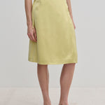 A person wearing a pale yellow knee-length Emmy Wrap Skirt by Herskind ApS, featuring an A-line silhouette tied at the waist with a simple side bow and black flat shoes.