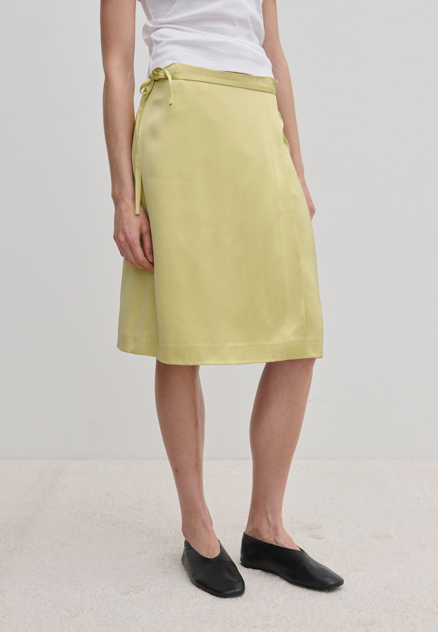 A person wearing a pale yellow knee-length Emmy Wrap Skirt by Herskind ApS, featuring an A-line silhouette tied at the waist with a simple side bow and black flat shoes.