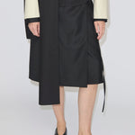 A person wearing a black, knee-length Emmy Wrap Skirt by Herskind ApS with an asymmetric hem, paired with a cream-colored jacket and black flat shoes.