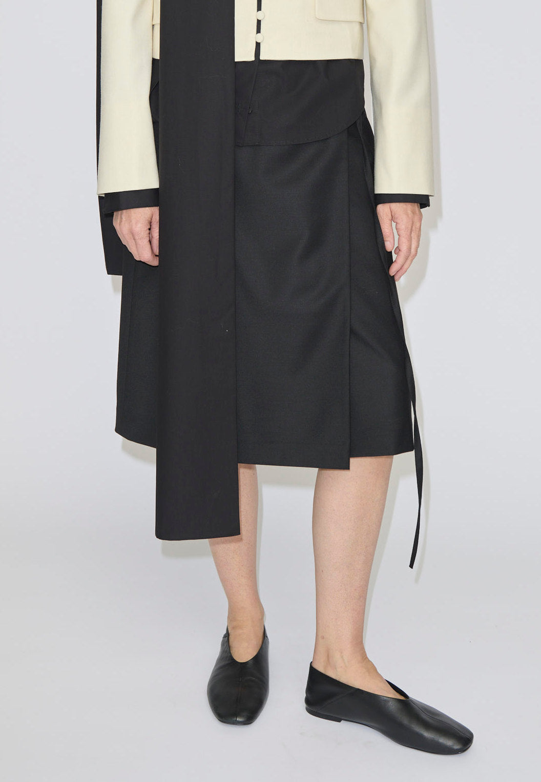 A person wearing a black, knee-length Emmy Wrap Skirt by Herskind ApS with an asymmetric hem, paired with a cream-colored jacket and black flat shoes.