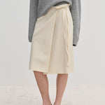 Cream-colored Emmy Wrap Skirt by Herskind ApS, a mid-waist wrap style with a side tie and A-line silhouette, worn with a grey sweater and black slingback flats.