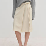Cream-colored Emmy Wrap Skirt by Herskind ApS, a mid-waist wrap style with a side tie and A-line silhouette, worn with a grey sweater and black slingback flats.