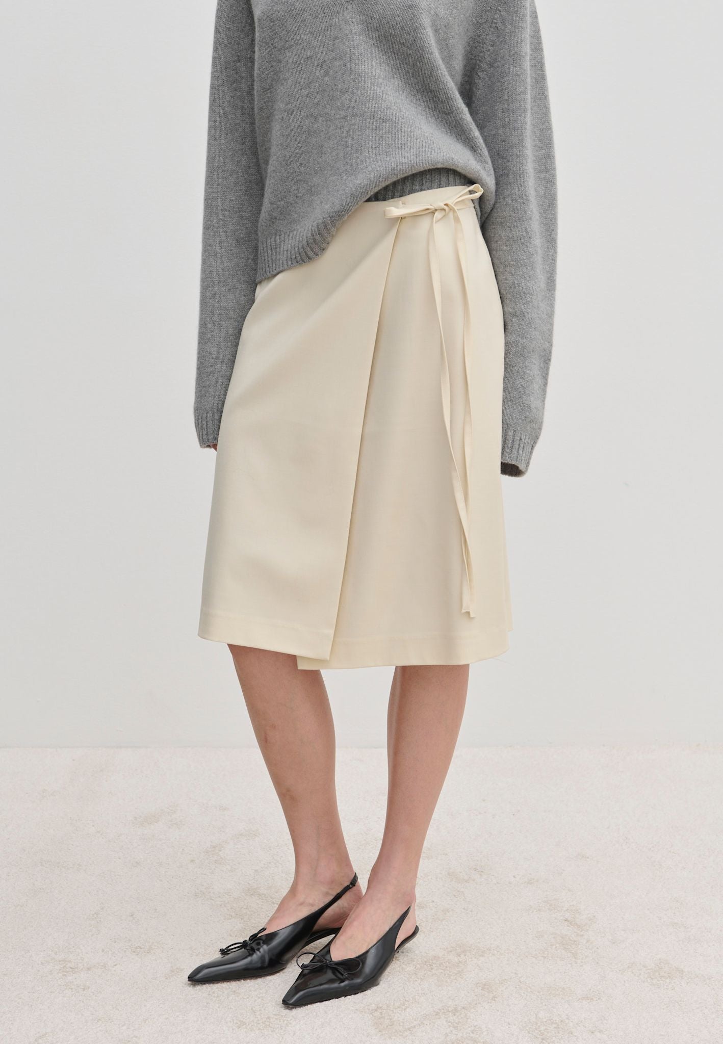 Cream-colored Emmy Wrap Skirt by Herskind ApS, a mid-waist wrap style with a side tie and A-line silhouette, worn with a grey sweater and black slingback flats.