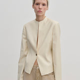 A person wears a cream-colored, fitted Henry Fitted Blazer by Herskind ApS with a stand collar, structured shoulders, and a single front button closure.