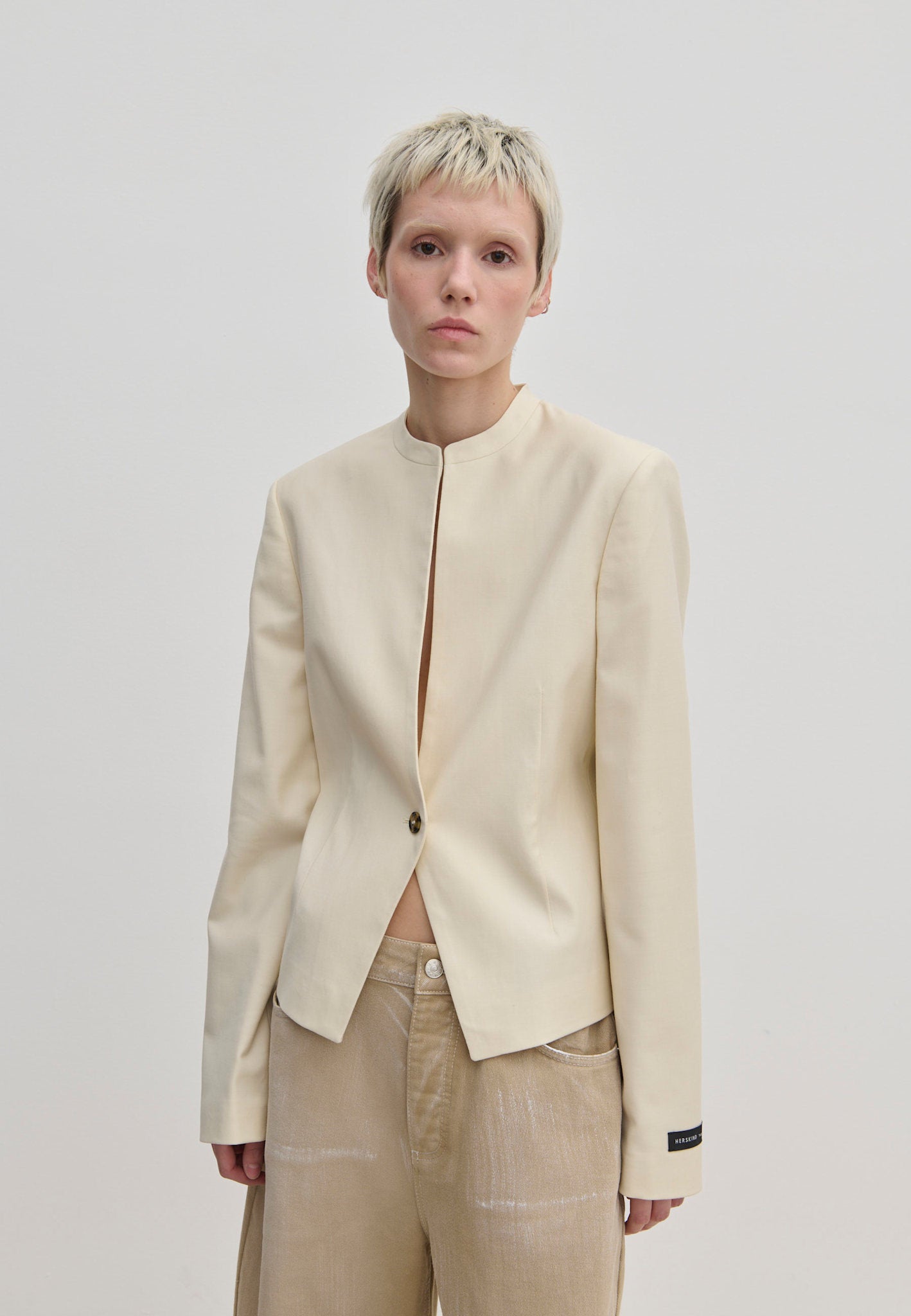 A person wears a cream-colored, fitted Henry Fitted Blazer by Herskind ApS with a stand collar, structured shoulders, and a single front button closure.