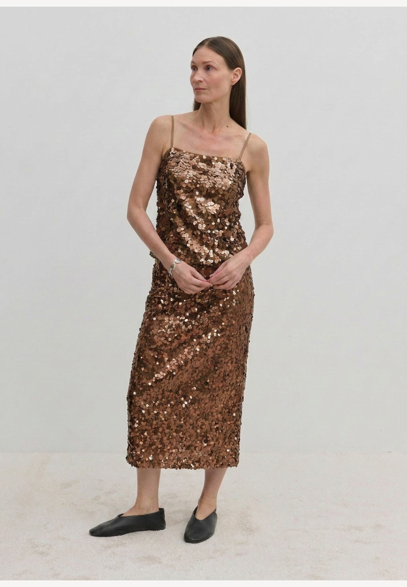 A person wearing a bronze sequined camisole top and the matching Anda Skirt, paired with black flat shoes.