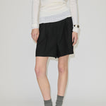 A person wearing a white long-sleeved top, black pleated Herskind ApS Reggy High Waist Shorts, grey socks, and black slip-on shoes.