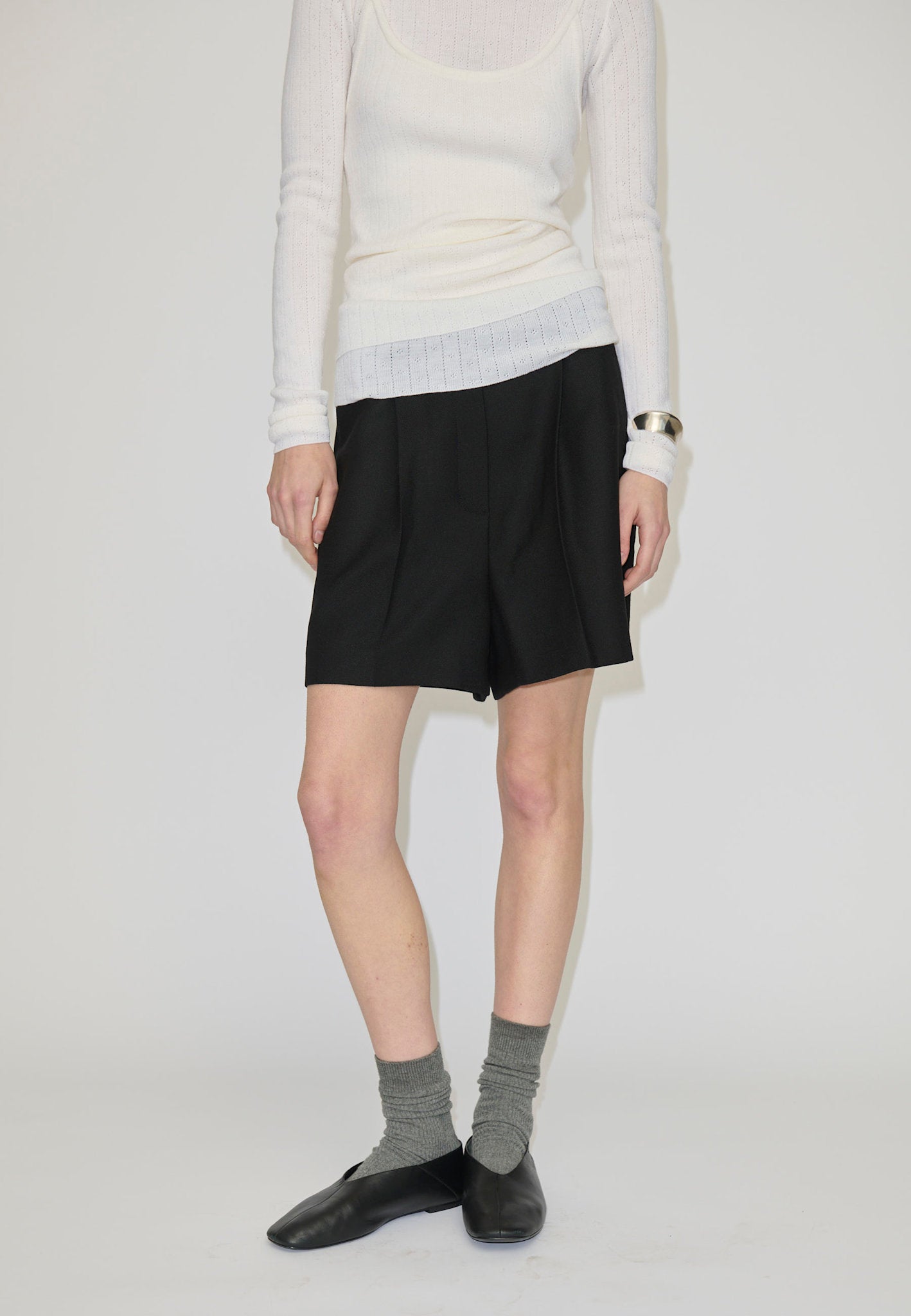 A person wearing a white long-sleeved top, black pleated Herskind ApS Reggy High Waist Shorts, grey socks, and black slip-on shoes.