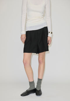A person wearing a white long-sleeved top, black pleated Herskind ApS Reggy High Waist Shorts, grey socks, and black slip-on shoes.