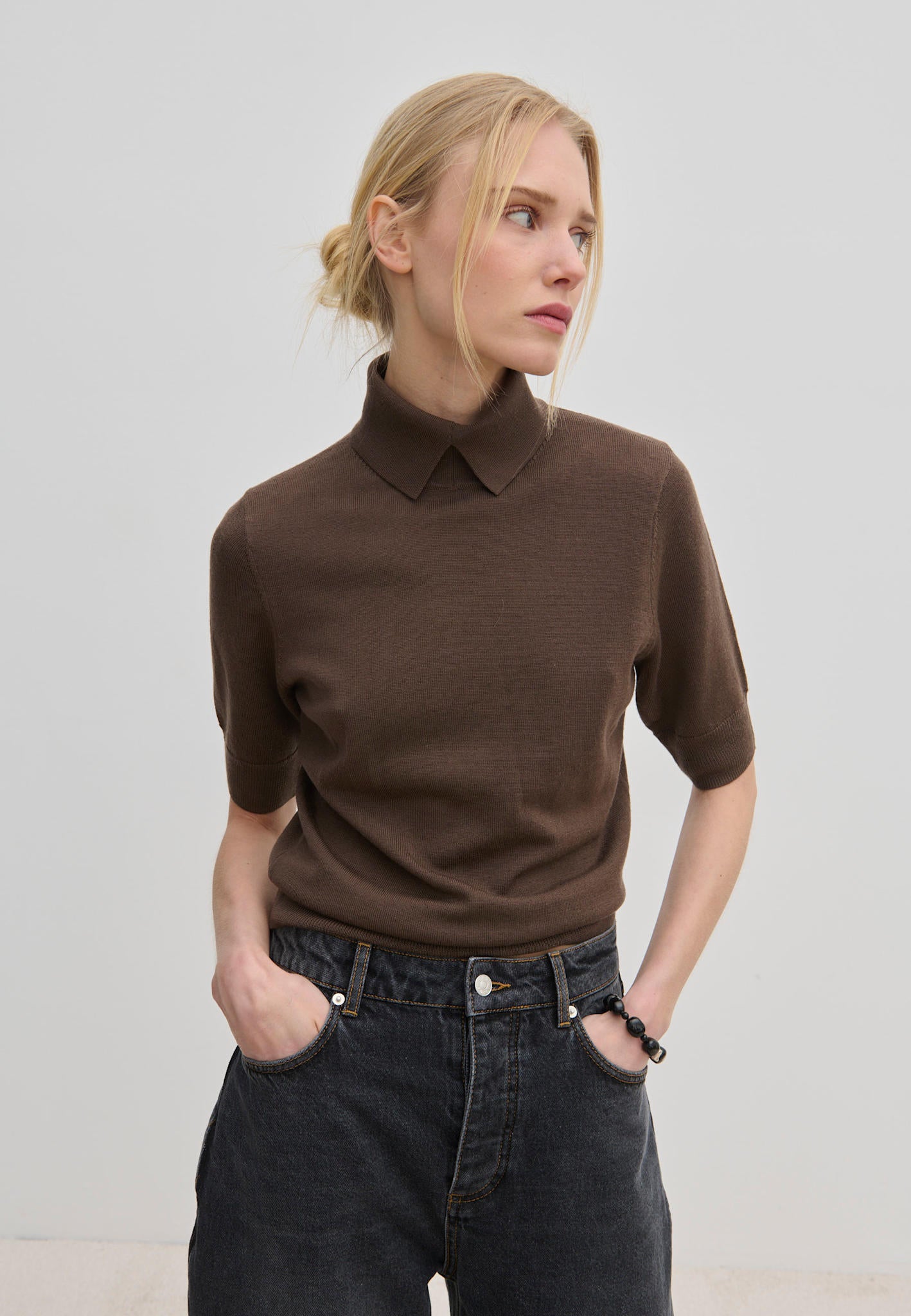 A person wears a short-sleeved brown merino wool Perry Knit Blouse by Herskind ApS with a collared neckline tucked into dark gray jeans.