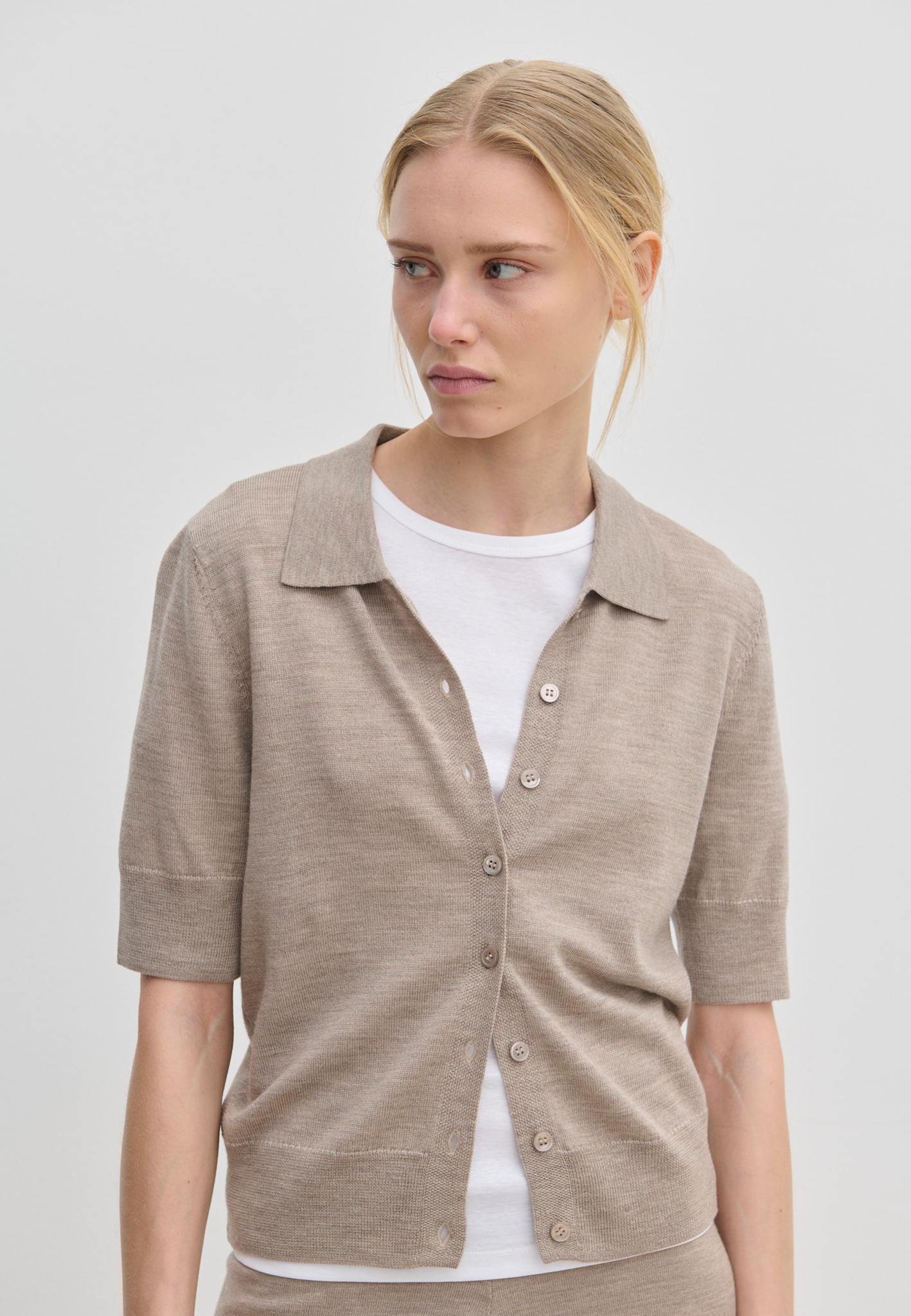 A beige, relaxed-fit Kaspar Knit Polo by Herskind ApS worn unbuttoned over a white t-shirt.