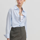 A woman wearing a light blue Lorenzo Shirt by Herskind ApS left open over a white tank top, paired with a dark grey skirt and a long beaded necklace.