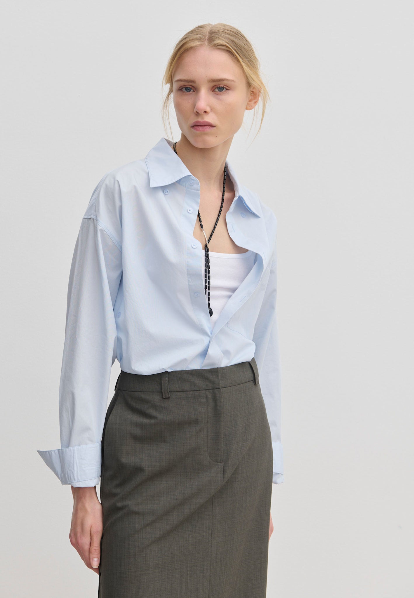 A woman wearing a light blue Lorenzo Shirt by Herskind ApS left open over a white tank top, paired with a dark grey skirt and a long beaded necklace.