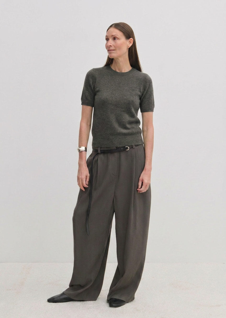 A woman wearing a charcoal grey Herskind ApS Thora Knit Blouse tucked into pleated wide-leg trousers with a thin black belt.