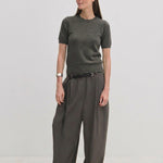 A woman wearing a charcoal grey Herskind ApS Thora Knit Blouse tucked into pleated wide-leg trousers with a thin black belt.