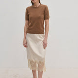A woman wearing a brown Thora Knit Blouse in wool and cashmere, a cream-colored midi skirt with a lace hem, and black heeled thong sandals.