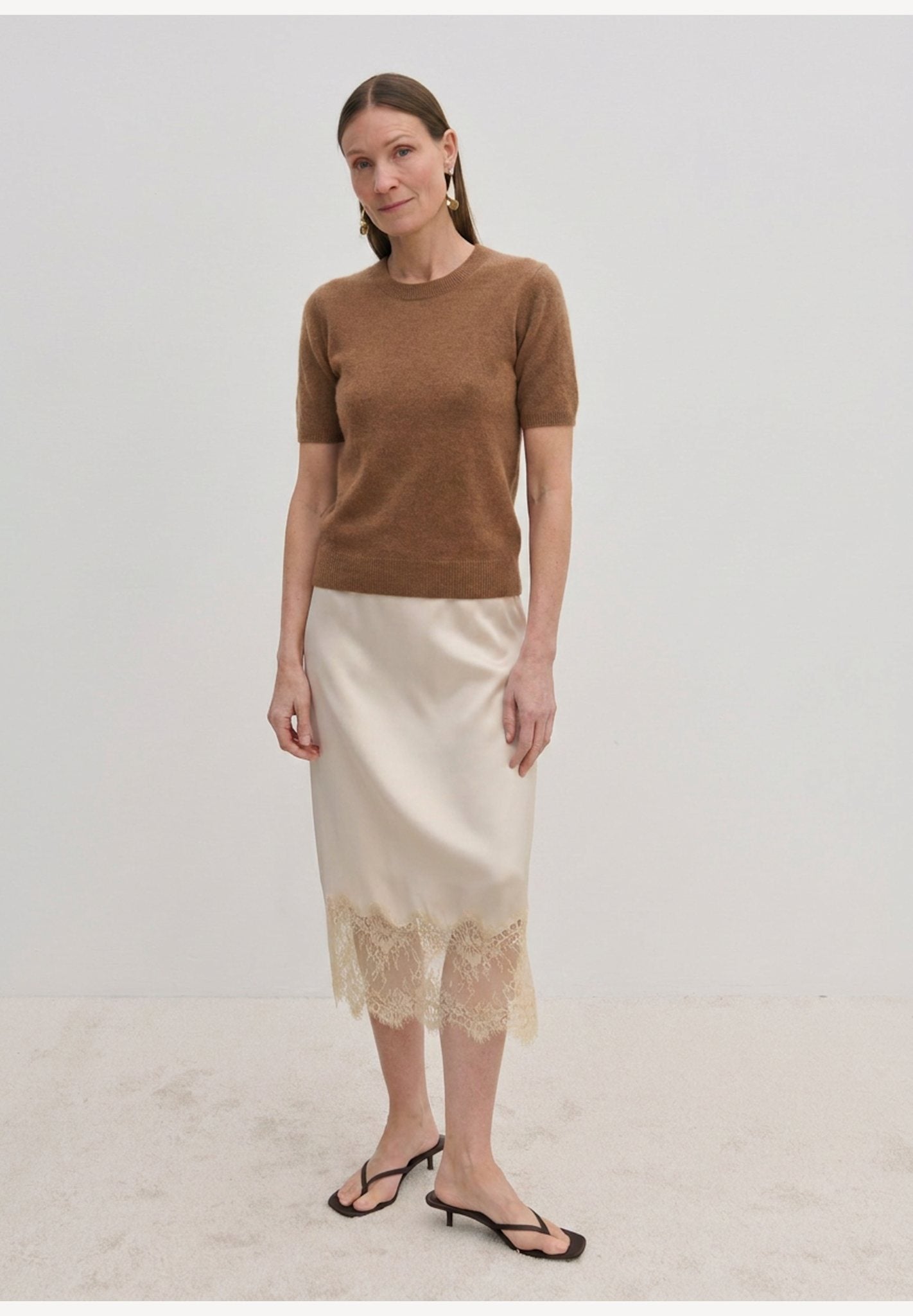 A woman wearing a brown Thora Knit Blouse in wool and cashmere, a cream-colored midi skirt with a lace hem, and black heeled thong sandals.
