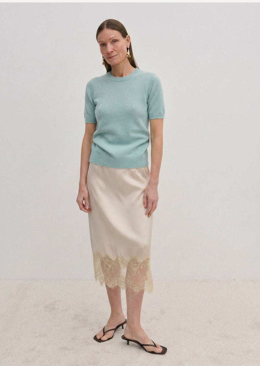 A woman wearing a pale green Thora Knit Blouse by Herskind ApS and a beige satin midi skirt with a lace hem, paired with black heeled sandals.