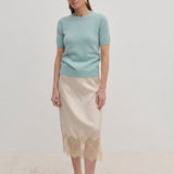 A woman wearing a pale green Thora Knit Blouse by Herskind ApS and a beige satin midi skirt with a lace hem, paired with black heeled sandals.