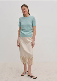 A woman wearing a pale green Thora Knit Blouse by Herskind ApS and a beige satin midi skirt with a lace hem, paired with black heeled sandals.
