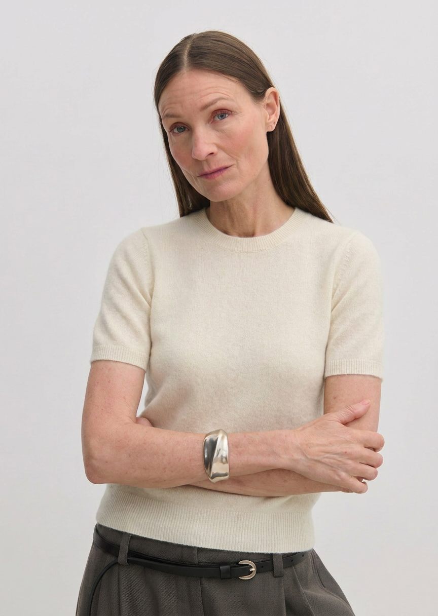 A woman wears a cream-colored, short-sleeved crew-neck Thora Knit Blouse by Herskind ApS, paired with grey trousers and a black belt.