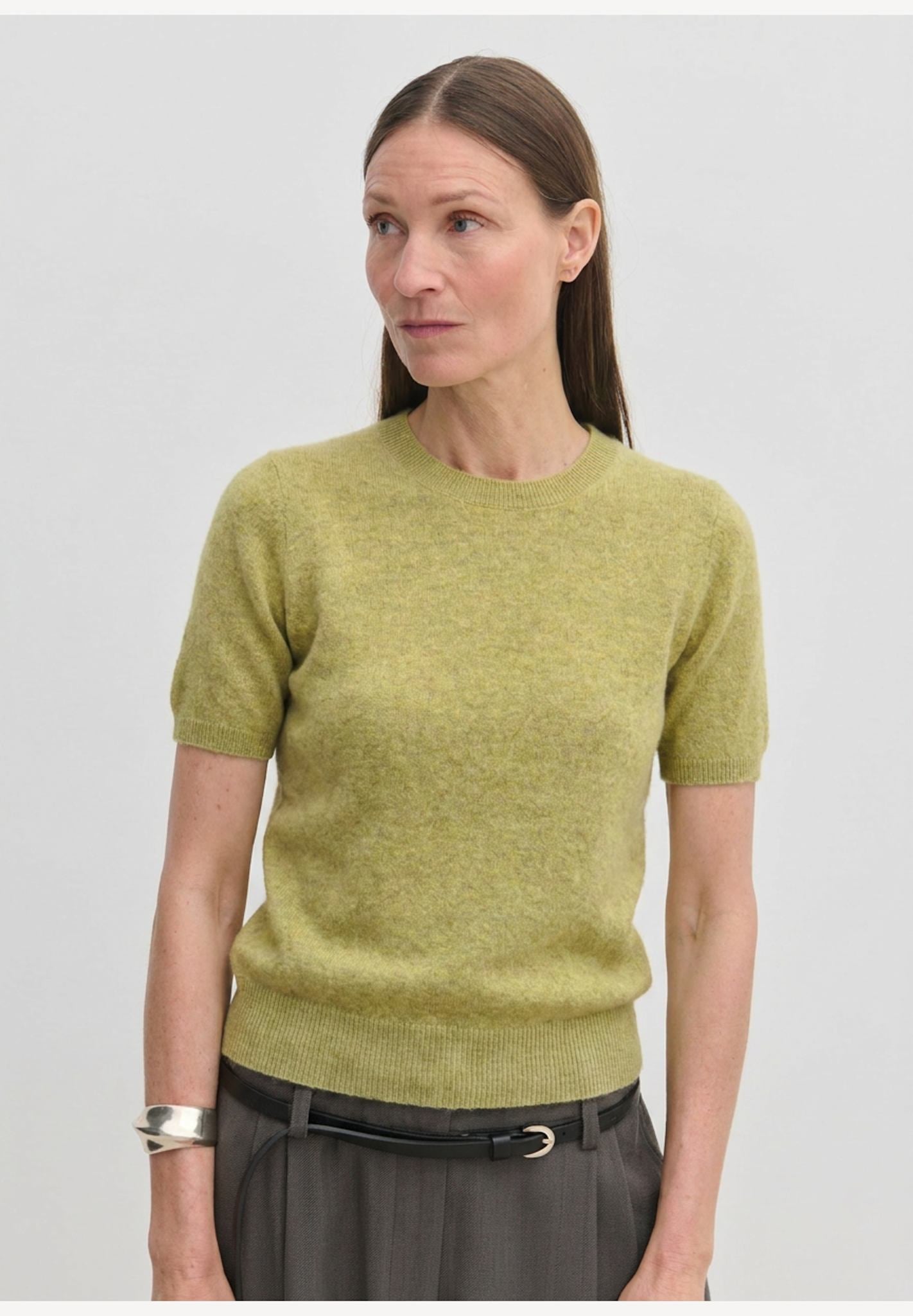 A woman wearing a lime green Thora Knit Blouse tucked into dark trousers with a thin belt.