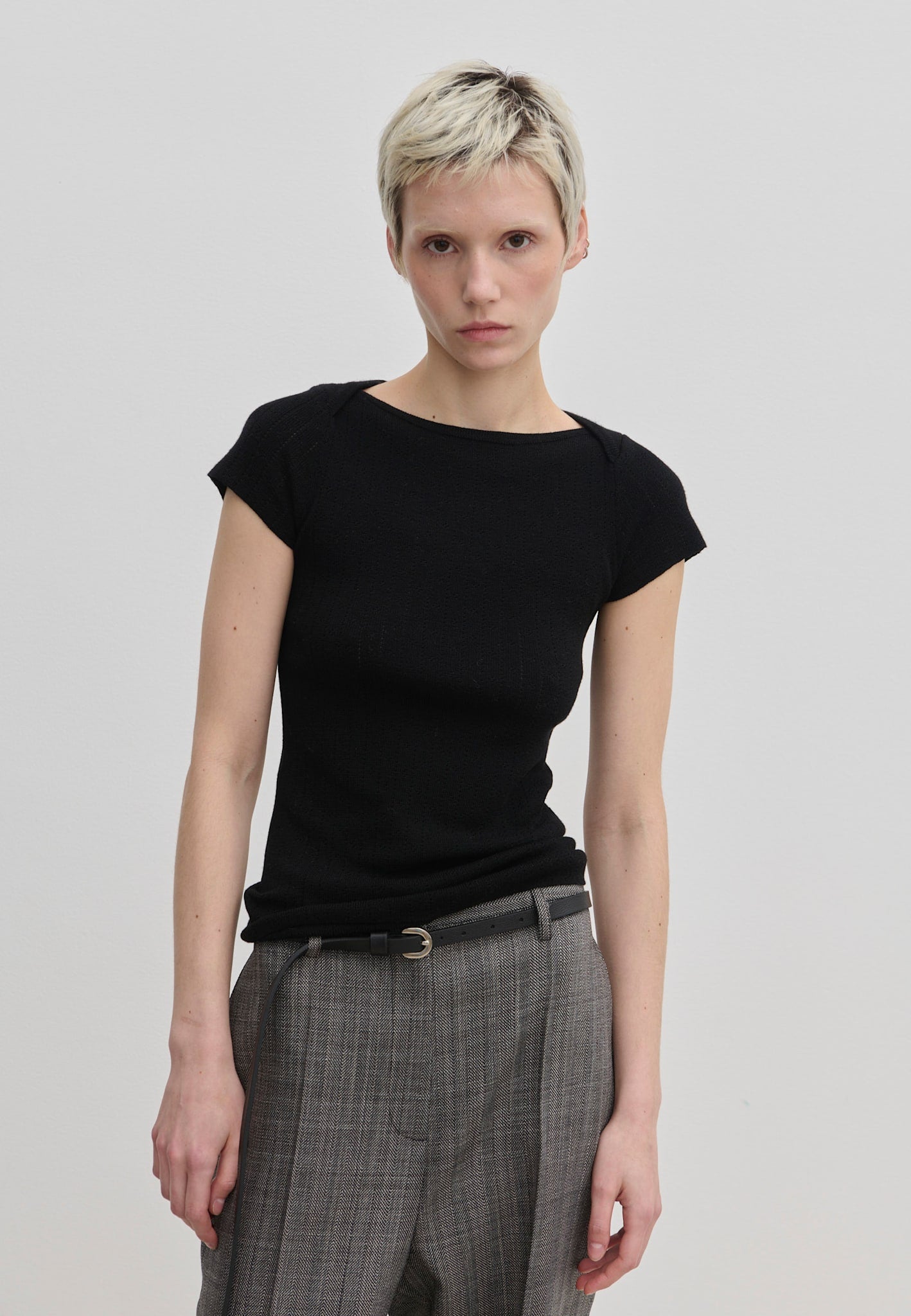 A person wearing a black, short-sleeved Erica Knit T-shirt by Herskind ApS, paired with grey patterned trousers and a thin belt.